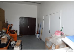 This photograph shows an interior room during renovation or construction work. The space contains white-painted walls, two doors (one dark wood, one white), and various construction materials, tools, and equipment scattered throughout, including an o