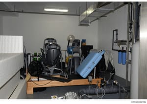 This is a photograph of a storage or utility room containing various exercise and fitness equipment, including exercise machines, equipment cases, water filtration systems, and miscellaneous storage items. The room appears to be part of an Epstein pr