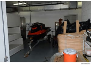 This photograph shows an interior garage or storage facility containing a red and black jet ski on a trailer, numerous wrapped packages and items covered in plastic sheeting, and various storage containers. The image appears to be evidence documentat