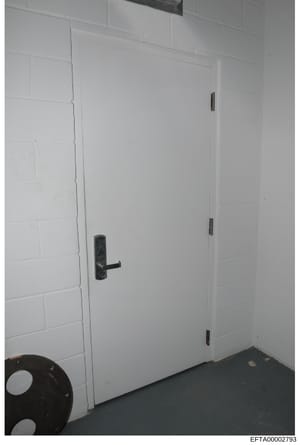 This is an evidence photograph showing the interior of a room with white painted brick walls and a white door with a dark metal handle. A black object (possibly a tire or circular item) is partially visible in the lower left corner. The image appears