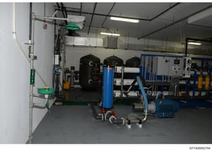 This photograph shows an industrial facility interior containing water treatment or filtration equipment, including large black pressure tanks, blue filtration units, a blue electric motor, piping systems, and control panels. The image appears to be 