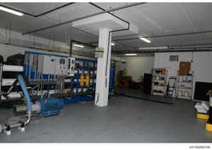 This photograph shows an industrial or mechanical equipment room, likely part of a property facility or basement. The space contains various blue and white industrial machinery, control panels, pumps, storage shelving, and utility equipment arranged 
