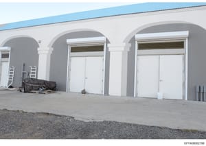 This is an evidence photograph showing the exterior of a white concrete building with distinctive arched architectural features and multiple garage doors. The image appears to be from an Epstein-related property investigation, documenting the structu