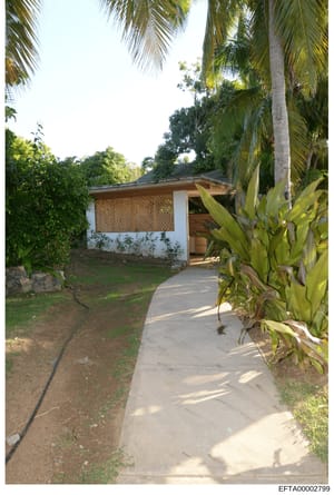 This photograph shows a small white building with latticed wooden panels and a covered entrance, situated within a tropical garden environment. The structure appears to be a guest house, storage facility, or ancillary building on a larger property, s