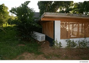 This is a photograph of an exterior structure, likely a guest house, pool house, or outbuilding on a property. The image shows a single-story white building with wooden lattice screening panels, surrounded by mature landscaping and tropical vegetatio
