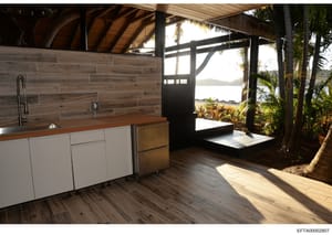 This is a photograph of a modern kitchen and open-air living area in a tropical beachfront property. The image shows a contemporary kitchen with white cabinetry and wood countertops, adjacent to a covered outdoor pavilion with views of water and dist