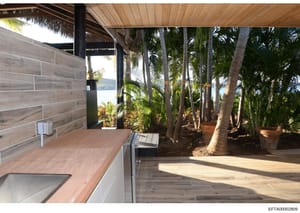 Photograph of an upscale waterfront property's covered outdoor living space, featuring a modern kitchen counter with wood finishes, thatched roof covering, and direct views of tropical landscaping and water beyond. The image documents the property's 