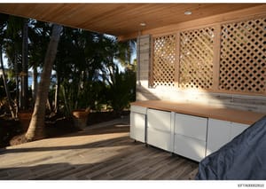 This photograph shows a covered outdoor patio area of a waterfront property, featuring a wooden ceiling, lattice privacy screens, built-in white cabinetry with wood countertop, and views of palm trees and water. The image appears to be part of proper