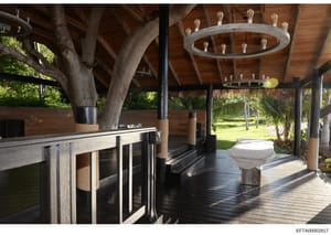 This photograph shows the interior of a covered outdoor pavilion or entertainment structure with exposed wooden beams, a kitchen counter with bar seating on the left, decorative circular light fixtures hanging from the ceiling, and views of landscape