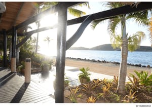 This is a photograph of a luxury beachfront property showing an interior covered patio or deck area with modern black beam construction, tropical landscaping, and direct views of a pristine beach and bay. The image appears to be documentation of real