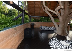 This is a photograph of an outdoor deck or patio space featuring modern design elements. The area includes wooden decking, a large natural tree trunk, custom seating with patterned cushions, and is surrounded by dense vegetation. The space appears to