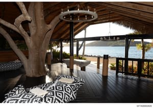 This is a photograph of an upscale beachfront property's covered outdoor deck or patio area. The image shows a modern, luxuriously appointed space with high-end finishes including a large circular chandelier, natural wood beam structural elements, de