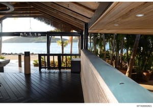 This is a photograph of a luxury beachfront property's interior deck and patio area. The image shows a covered wooden structure with expansive ocean views, tropical landscaping with palm trees, modern architectural design including wooden beam ceilin
