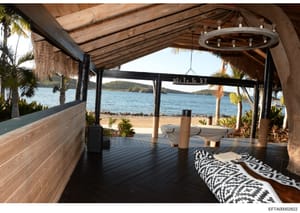 Photograph of a luxury beachfront pavilion or covered outdoor living space featuring modern rustic design with exposed wooden beams, decorative circular chandelier, and contemporary furnishings overlooking a scenic bay with palm trees and distant isl
