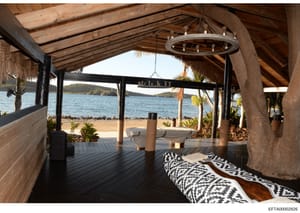 This photograph shows an interior view of a covered beachfront pavilion or cabana structure with exposed wooden beams, a decorative chandelier, and modern furnishings. The space overlooks a sandy beach, calm ocean waters, and distant forested islands