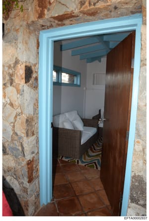 This is a photograph of a small interior room or space accessed through a bright blue-framed doorway in a weathered stone building. The room contains a rattan chair with white cushions and a dark wooden door. The image appears to be documentation of 