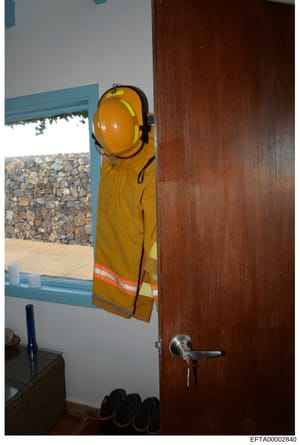This is an evidence photograph showing the interior of a room with a window overlooking a stone courtyard or exterior area. A yellow hard hat and safety vest hang on the wall beside a dark wooden door. The image appears to be documentation of a prope
