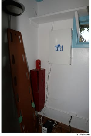 This is a photograph of an interior room space showing a wall-mounted white first aid kit, emergency equipment including a red fire extinguisher, a brown stretcher or medical transport device, and various electrical outlets and wiring. The photo appe