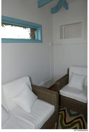 This is an evidence photograph showing a small bedroom interior with two twin beds with white linens and wicker headboards. The room features turquoise-trimmed windows and architectural elements, white walls, and minimal furnishings. The photograph a