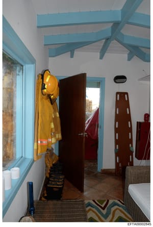 This photograph shows an interior room with distinctive architectural features including a turquoise/blue painted beam ceiling and trim. The room contains safety and emergency equipment including a yellow firefighter or rescue suit hanging on the wal