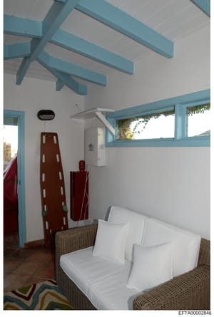 This is a photograph of an interior room featuring modern coastal-style design with turquoise and white color scheme. The room contains a wicker seating area with white cushions, exposed beam ceiling details, and large windows with ocean views. The i