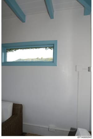 This is a photograph of an interior room, likely a bedroom or guest room, showing architectural details including turquoise-painted ceiling beams and window frames, white walls, and a horizontal window with a landscape view. The image appears to be e
