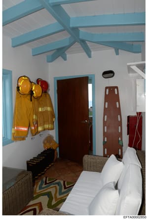 This photograph shows an interior room with distinctive architectural features including turquoise exposed beam ceiling, white walls, and a decorative geometric patterned rug. The room contains emergency and safety equipment including firefighting ge