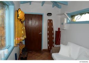 This photograph shows an interior room with turquoise trim and white walls, containing various utility items including firefighting equipment (yellow protective gear and helmet), a brown ladder, red fire extinguisher, ironing board, white bedding/fur