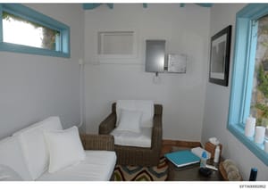 This is an interior photograph of a residential room featuring wicker seating furniture, white pillows, and basic furnishings. The room has turquoise-trimmed windows and doors, white walls, and appears to be a modest living or sitting area. The photo