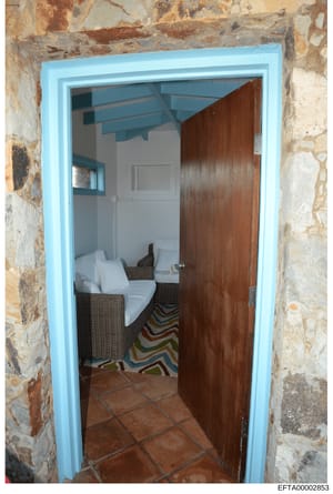 This is a photograph of an interior room, likely a bedroom or guest room, showing a doorway framed by weathered stone walls with a bright blue trim. The room contains wicker furniture with white cushions and a colorful striped rug. The image appears 