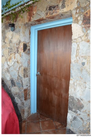 This photograph shows an interior doorway with rustic stone walls featuring multi-colored stones in beige, tan, and brown tones. The wooden door has a dark brown finish and is framed with bright blue trim. A small black fixture is visible on the left