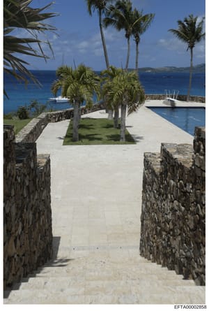 This photograph shows a luxurious waterfront property featuring a manicured courtyard with palm trees, an infinity pool, and a clear view of the turquoise Caribbean Sea with moored vessels. The stone-walled pathways and high-end construction suggest 