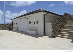This is a photograph of a modern, single-story white building with a flat roof and dark trim, featuring a white garage door and stone retaining wall. The structure appears to be a utility or service building on a residential or estate property, photo