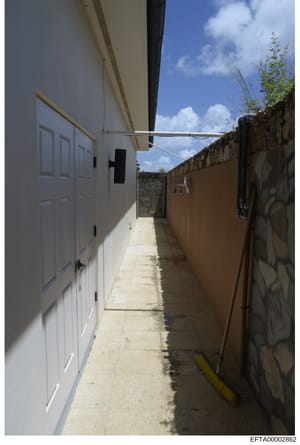 This photograph depicts a narrow outdoor alleyway or service corridor between two buildings. The left side shows a modern white building with closed doors, while the right side features older stone/brick walls. The space appears to be a property serv