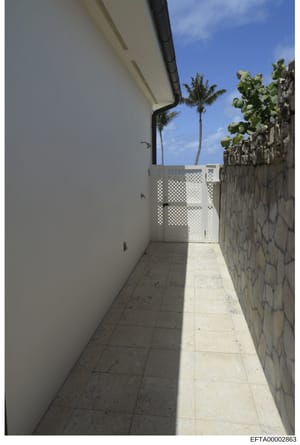 This photograph shows a narrow, covered walkway or passage at what appears to be a tropical residential property. The image captures a tiled pathway between a white wall and a stone boundary wall, with a white lattice gate visible in the distance and