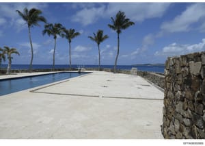 This is a photograph of a luxury oceanfront property featuring a swimming pool, expansive terrace with ocean views, and palm trees. The image shows a high-end residential or hospitality property with Caribbean-style architecture and direct beach acce
