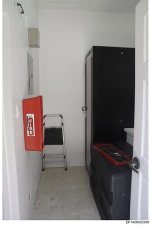 This is a photograph of a narrow interior room or hallway containing utility and safety equipment. The space shows a red fire extinguisher mounted on the left wall, a metal step ladder in the center, and a large black cabinet or equipment enclosure o