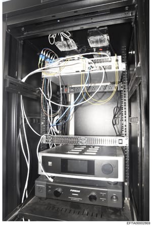 This is a photograph of a professional server rack containing networking and audio/visual equipment. The image shows multiple devices including cooling fans, network switches, amplifiers, and cabling infrastructure. This type of equipment documentati