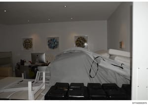 This is a photograph of a modern bedroom interior, showing a bed with white linens, contemporary wall art, recessed lighting, and office/workspace elements. The image appears to be evidence documentation from the investigation, likely taken during a 