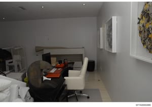 This photograph shows the interior of a residential bedroom or suite, featuring a bed, workspace with an orange/red desk, white chair, and wall-mounted artwork. The room appears to be part of a property under investigation, documented as evidence in 