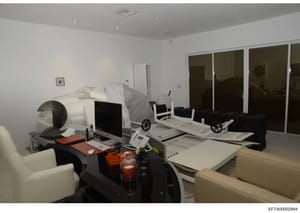 This is an evidence photograph depicting a modern residential bedroom that has been converted into a multi-functional space with office equipment, exercise machines, and medical devices. The room shows a bed, computer workstation, massage or medical 