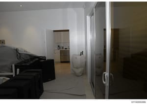 This is a photograph of an interior residential space showing a modern, minimalist apartment or condo unit. The image captures a combined living/kitchen area with light-colored walls, tile flooring, black furniture, and modern fixtures. The photo app