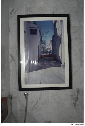 This is a photograph of a framed artwork depicting a narrow European street with white-washed buildings, bare trees, and clear blue sky. The photograph shows the framed print hanging on a marble-tiled wall in what appears to be a bathroom, with a sho