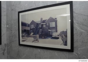 This is a photograph of a framed image showing the exterior of a brick residential property with multiple chimneys, external stairs, windows, and a parked vehicle in the driveway. The framed photograph is mounted on a marble-tiled wall. The image app