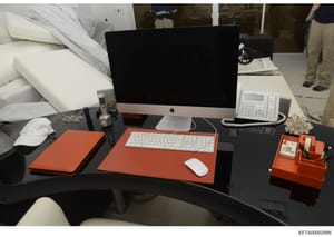 This photograph shows a professional office desk arrangement featuring a modern Apple iMac computer, white keyboard and mouse, and various orange-colored desk accessories including a mousepad, organizer, and notebook. The desk also includes a white t