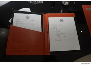 This photograph shows an open leather portfolio containing two pages of handwritten notes with what appears to be an official seal or emblem at the top of each page. The left page contains handwritten text in what may be a Cyrillic or non-Latin scrip