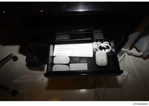 This is an evidence photograph showing a black desk organizer containing computer equipment including a white keyboard, mouse, remote control, power adapter, cables, and other electronic accessories. The photo appears to be part of a physical evidenc