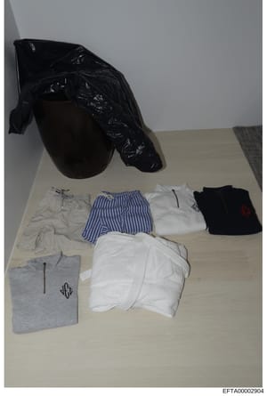 This is an evidence photograph showing clothing items laid out on a light-colored surface, including underwear, striped shorts, t-shirts with monograms, and a black plastic bag. The items appear to be catalogued evidence from a search or seizure cond