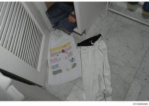 This is an overhead photograph of clothing and personal items laid out on a marble floor, including jeans, a white shirt with colored drawings, white pants, and a black garment. The photo appears to be evidence documentation from an investigation, sh