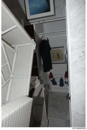 This is an evidence photograph showing the interior of a closet or small dressing room, featuring white shelving with folded linens or textiles, hanging clothing (including a black garment), footwear on lower shelves, and what appears to be a framed 